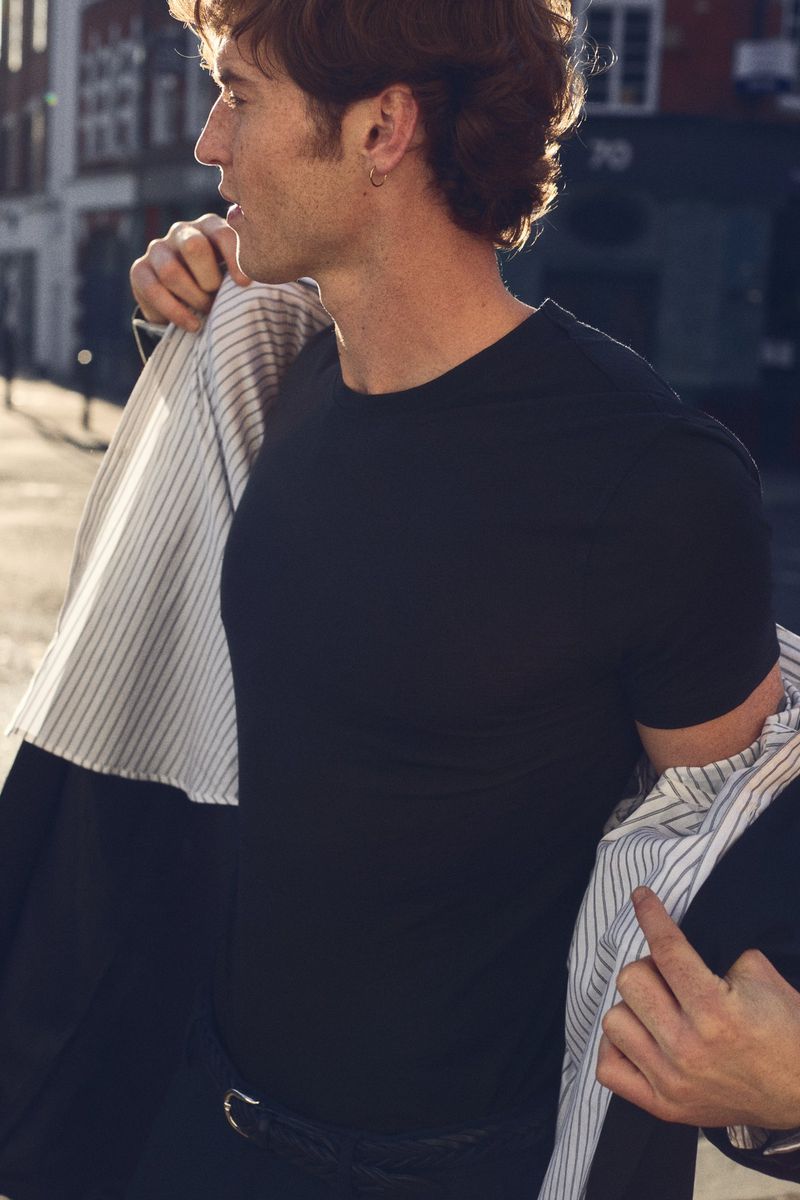 Adult male in black t-shirt and striped blazer stands outdoors on city street on sunny day looking left in medium studio shot.