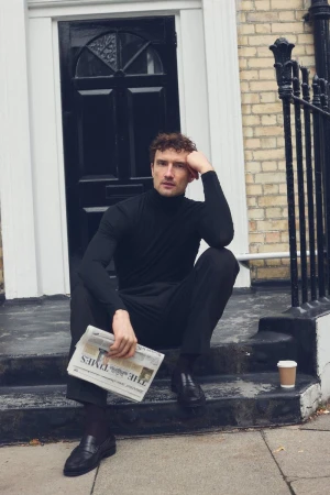 Model Jacob Coupe wearing a black turtleneck and trousers, sitting on stone steps holding a newspaper outside a residential building.
