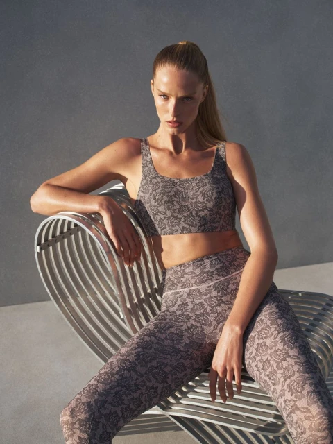 A woman in fitness apparel with a floral pattern sits on a metal chair in a brightly lit setting with a plain background.