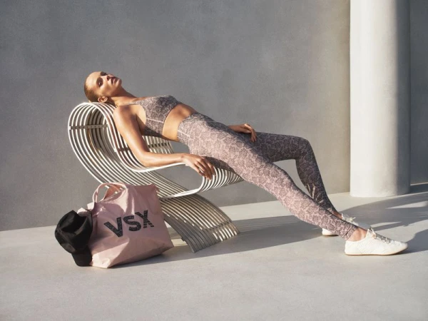 Model reclining on a white wire lounge chair wearing patterned athletic wear alongside a pink tote bag.