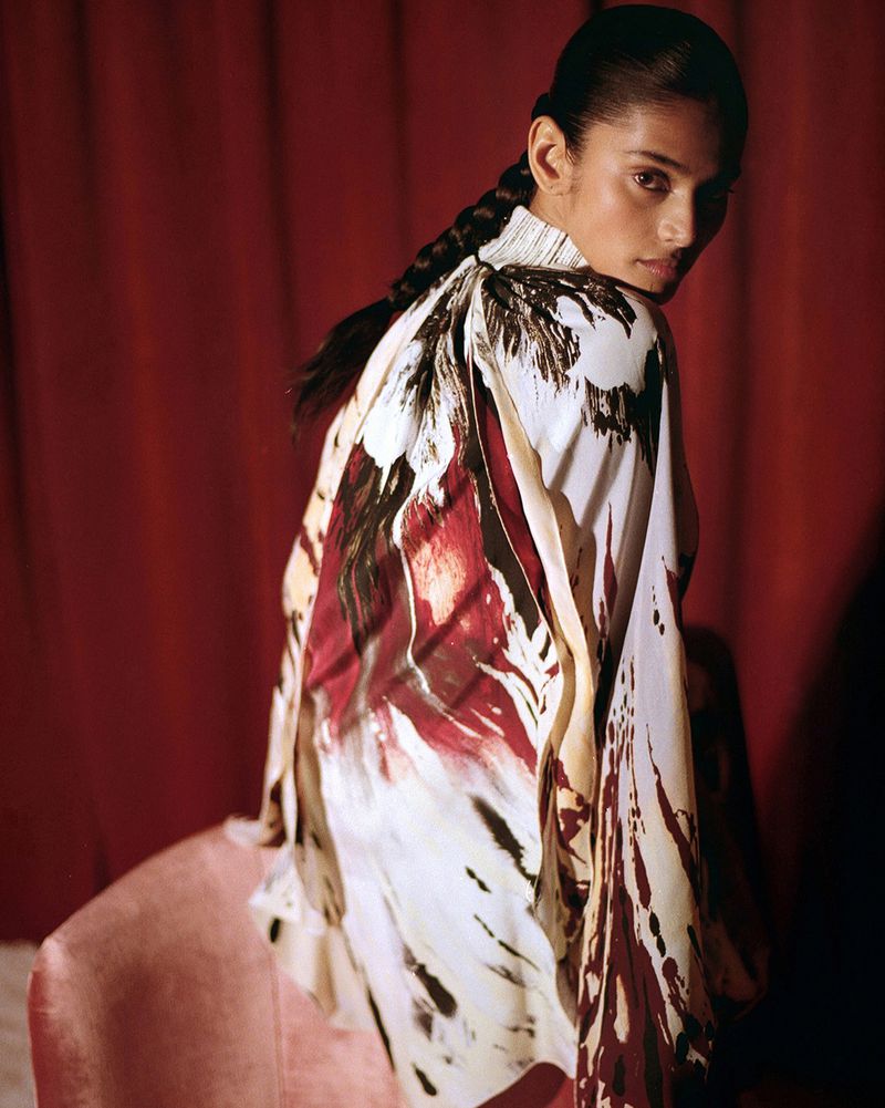 Adult female with braided hair wearing a patterned white and red long-sleeve top in a dimly lit indoor setting with a red backdrop.