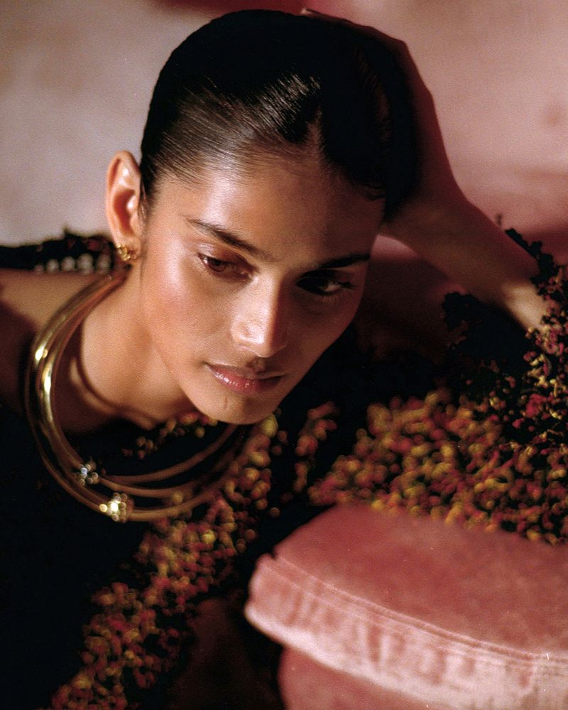 Portrait of an adult female model with dark hair, wearing gold jewelry, laying on a pink pillow in a studio setting.