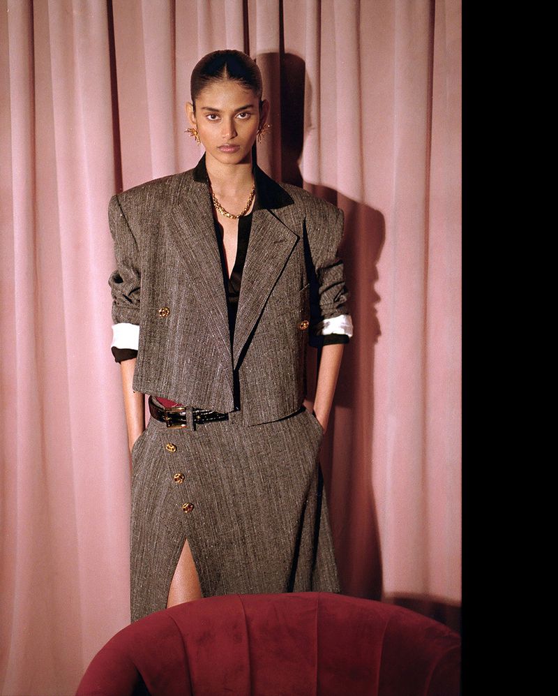 A sophisticated fashion portrait of an adult female wearing a grey tweed suit in front of a pink curtain backdrop, creating an editorial look.