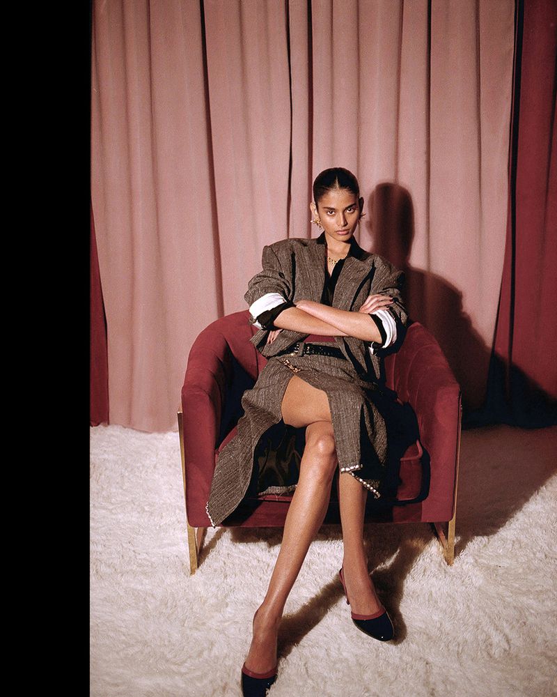 Woman in oversized blazer sits in red velvet chair in studio with pink drapes and white carpet, looking at the camera with arms crossed.