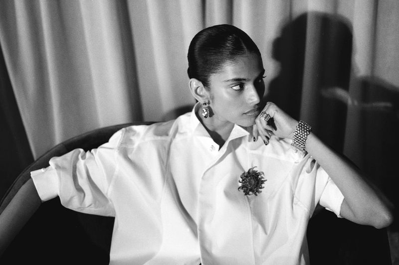 Monochrome portrait of a stylish adult female in white shirt, with jewelry, looking left against a curtain and shadow backdrop.