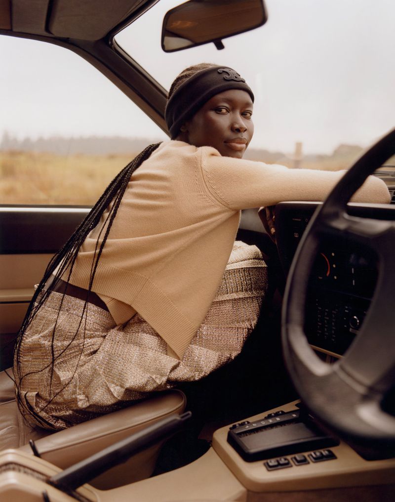 Woman in vintage car wearing beige sweater, metallic skirt and black headband looking at camera, with rural background, outdoor shot.