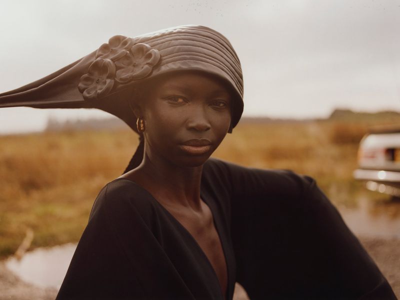 Elegant portrait of a black woman wearing a large black hat with floral embellishments, posing outdoors in a field by a car.