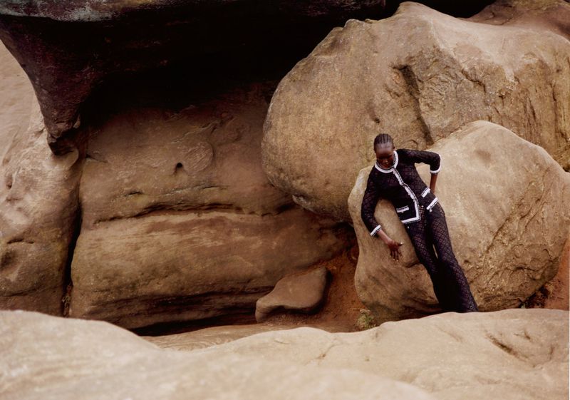 An adult woman poses in a fashionable black lace suit with white trim, surrounded by large boulders in an outdoor, rocky environment.