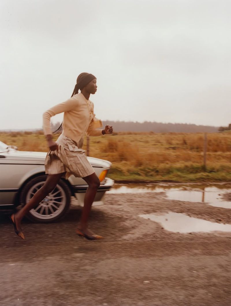 Young black woman in motion, outdoors. She is running on an overcast day in casual clothing alongside a parked silver car.