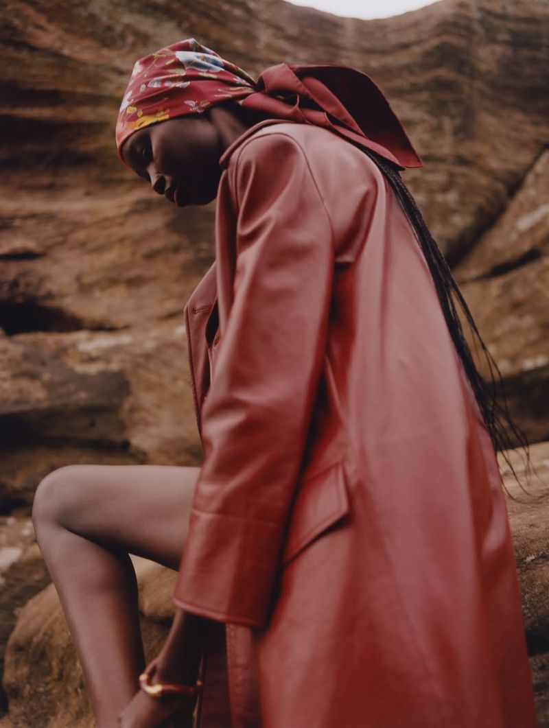 Fashion shot of a Black woman wearing a red coat and headscarf, sitting on rocks in an outdoor setting, capturing a blend of elegance and nature.