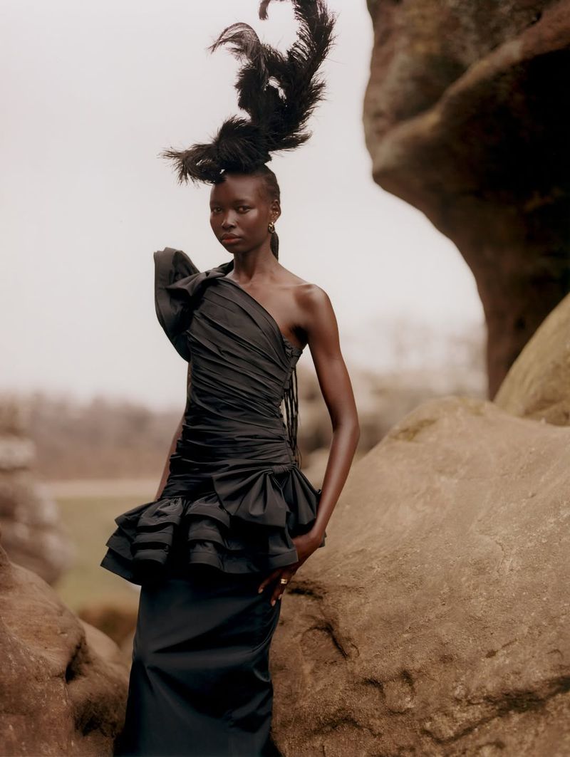 A statuesque Black woman poses in a dramatic black dress and feathery headpiece against a rock formation in a fashion editorial.