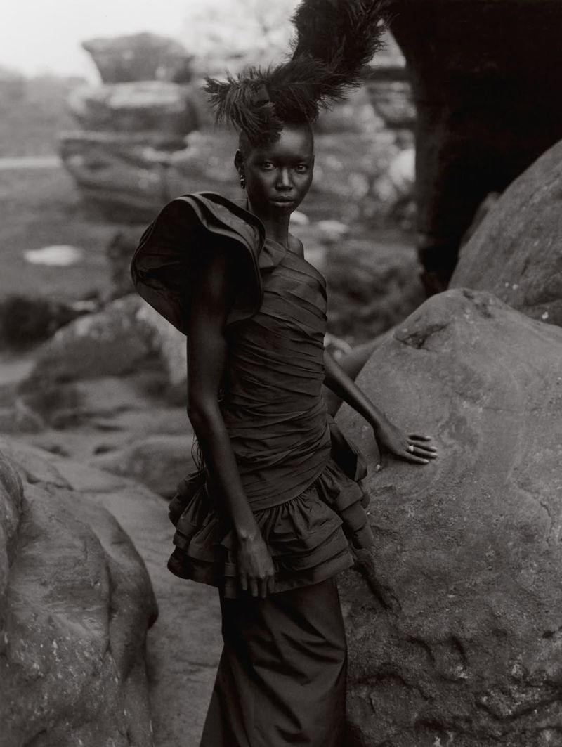A solitary, dark-skinned woman poses amongst rock formations wearing a formal dress and feathered headpiece for a fashion editorial.