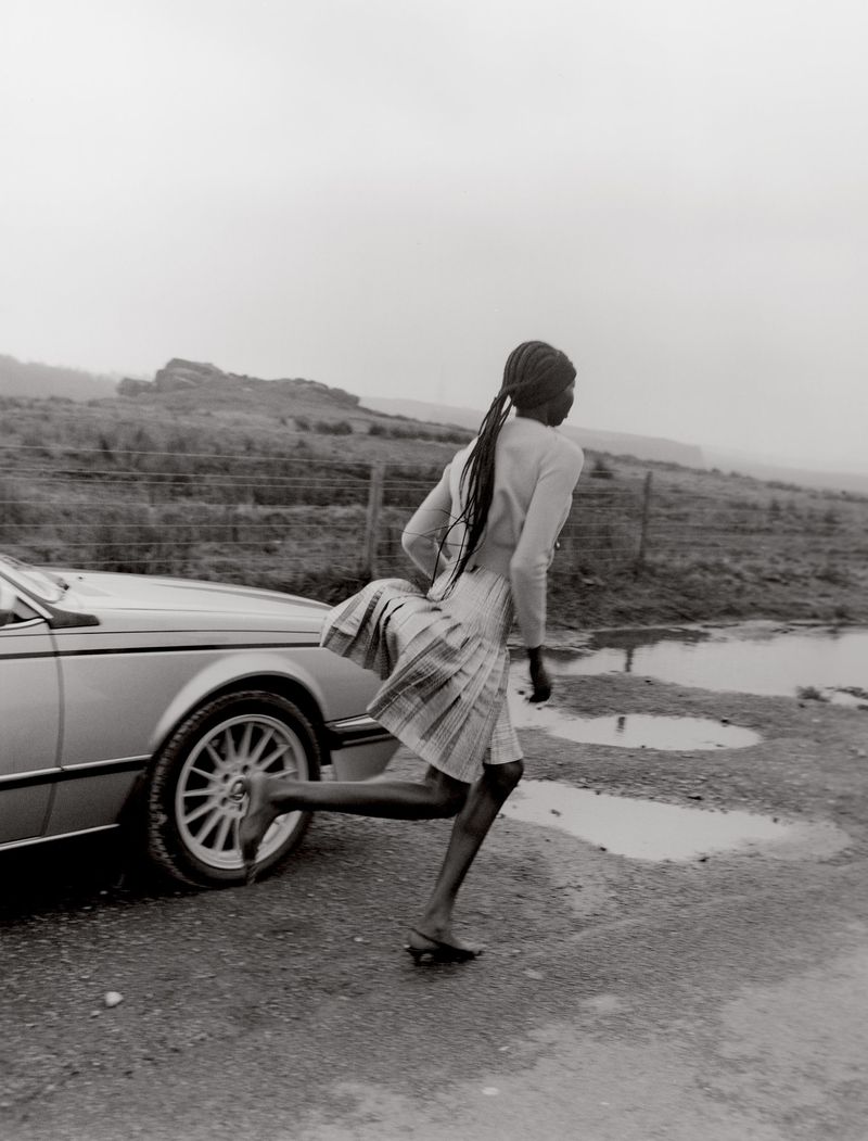 Monochrome shot of a Black woman running next to a vintage silver car in a rural setting with puddles after a rain.