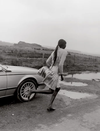 Monochrome shot of a Black woman running next to a vintage silver car in a rural setting with puddles after a rain.