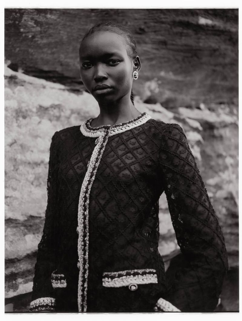 A single African woman stands in front of a light stone surface wearing a black jacket with white trim, looking forward in a close shot.