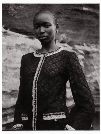 A single African woman stands in front of a light stone surface wearing a black jacket with white trim, looking forward in a close shot.