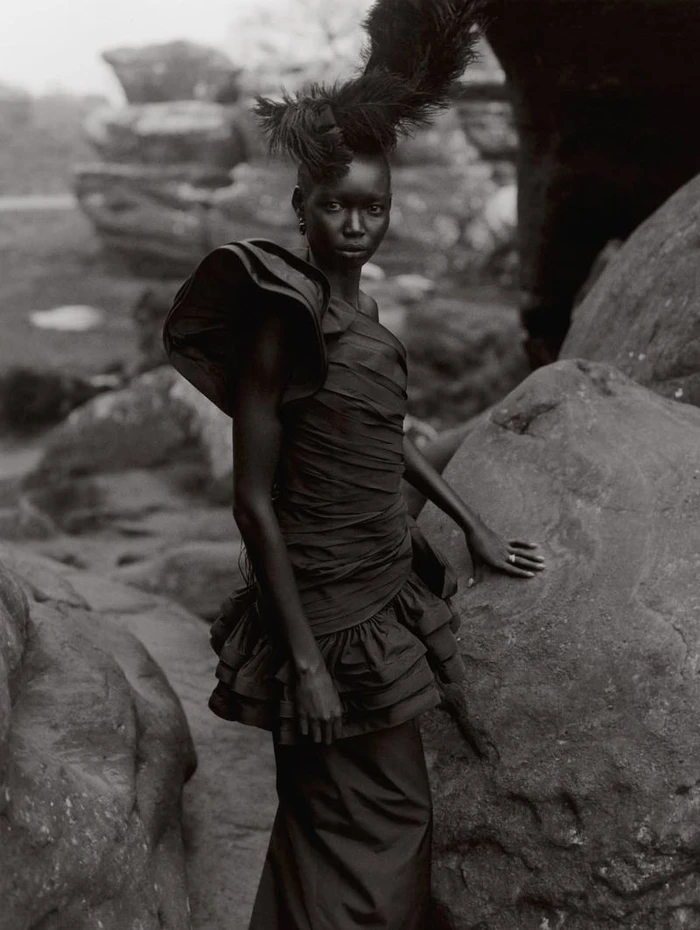 Model Amam Abit in a black ruffled dress with feather headdress standing among rocky boulders outdoors in a monochrome fashion portrait