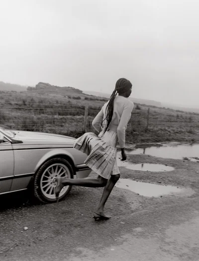 Model with long braided hair running past a vintage car on a wet gravel road under an overcast sky.
