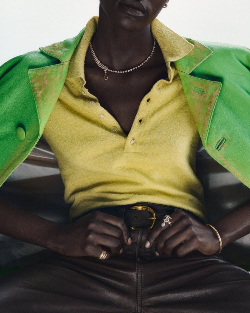 A woman with dark skin wearing a yellow polo shirt, green jacket, jewelry, and leather pants while seated indoors.