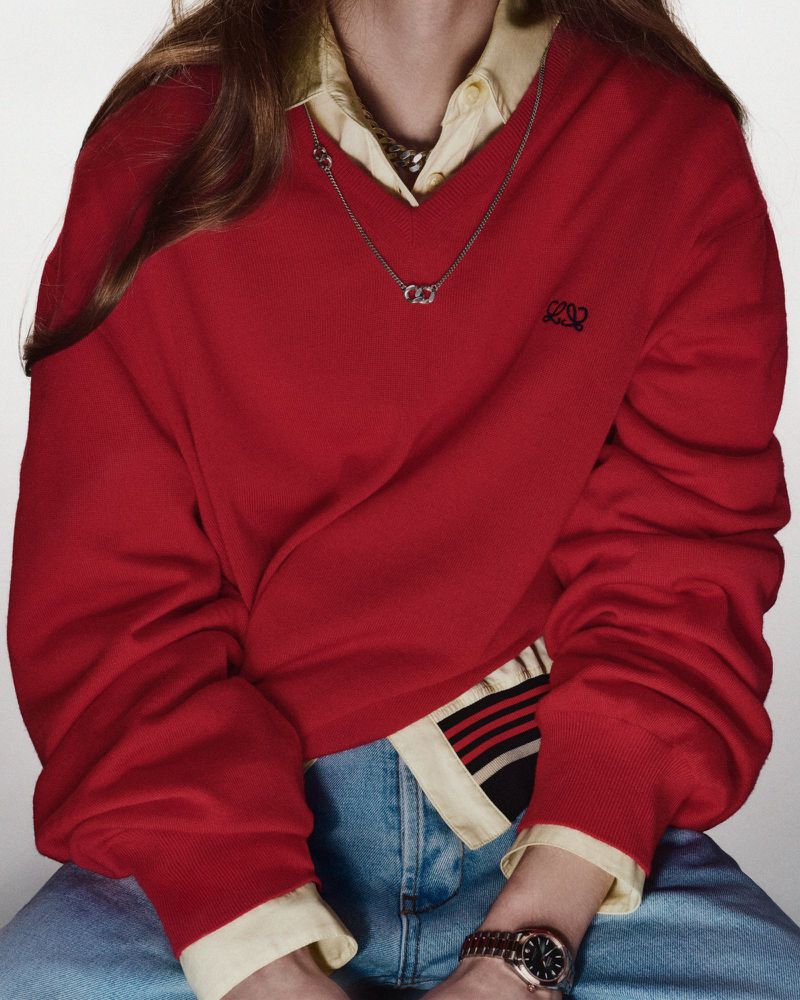Close-up of a person wearing a red v-neck sweater over a collared shirt with blue jeans and a watch, posing indoors.