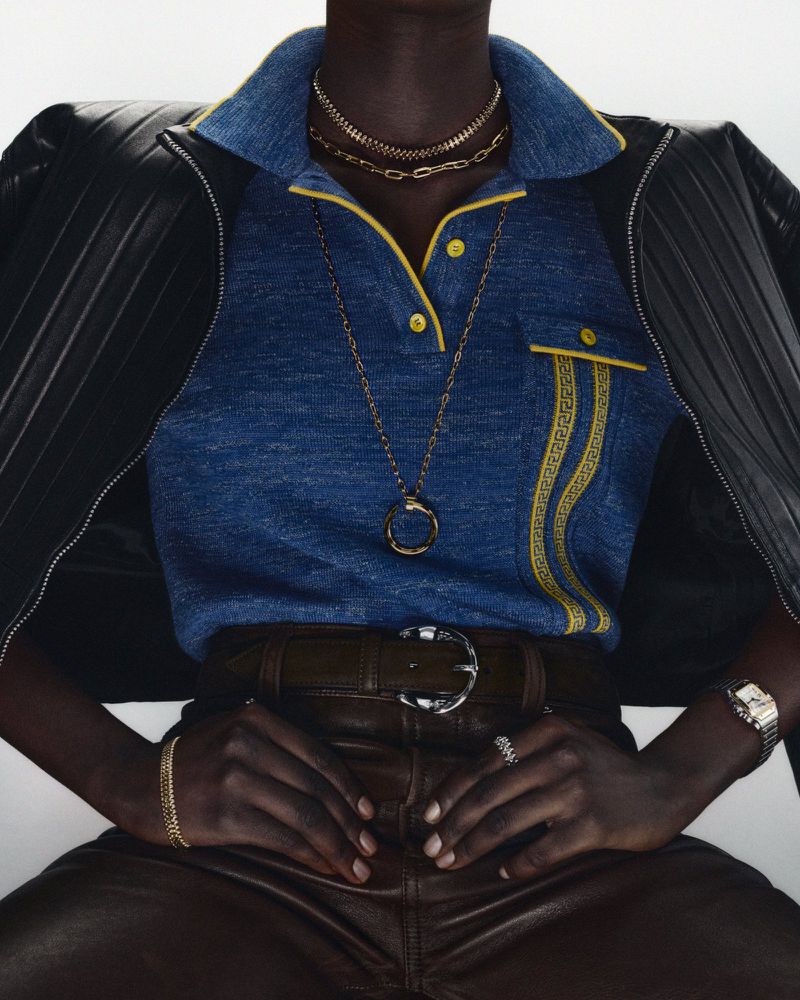 Dark-skinned model sits wearing a blue polo shirt, brown leather pants, and a black leather jacket, adorned with gold jewelry.