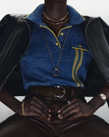 Dark-skinned model sits wearing a blue polo shirt, brown leather pants, and a black leather jacket, adorned with gold jewelry.
