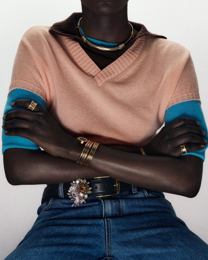 Dark-skinned model poses with arms crossed wearing a pink v-neck sweater, blue jeans, and gold jewelry in a fashion editorial studio shot.