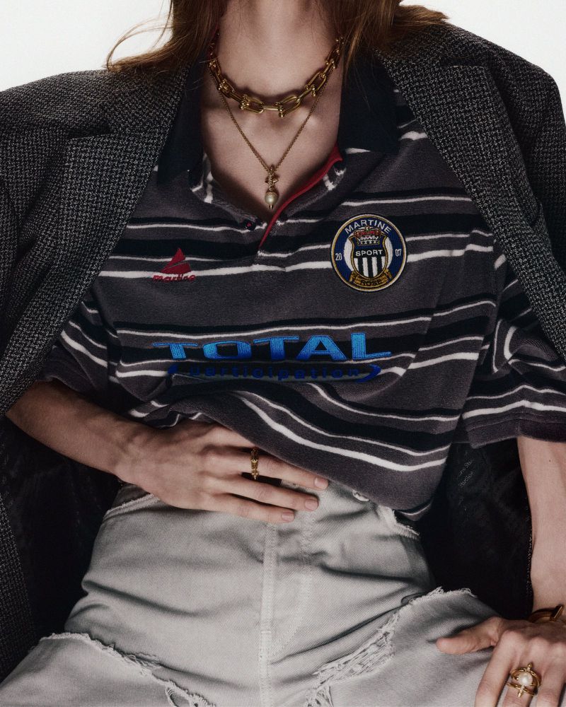 Close-up of an adult with gold necklaces, wearing a striped polo and grey blazer in an indoor setting with neutral background.