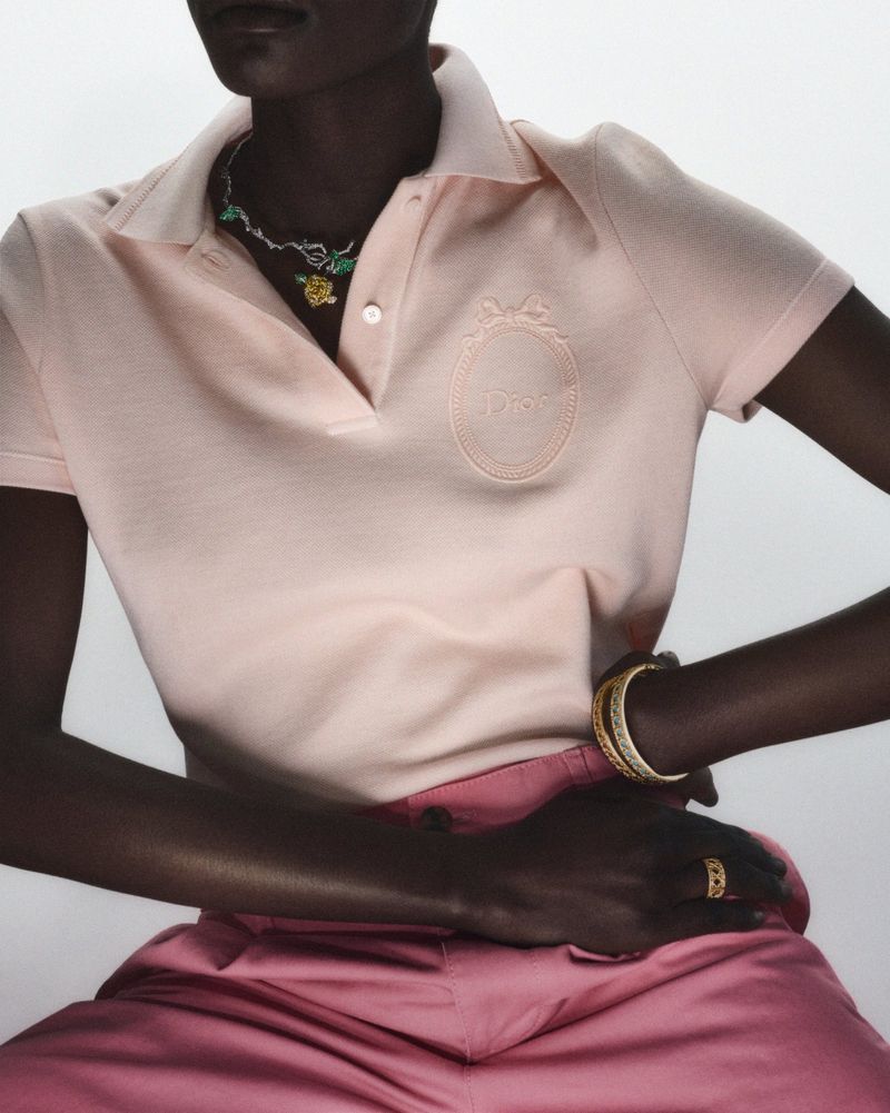 Model wearing a light pink Dior polo shirt and pink pants, accessorized with gold bracelets and a necklace, posing in a studio setting.