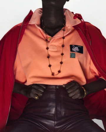 Person in orange polo shirt and leather pants with a red jacket, gold necklace, and rings against a plain white studio backdrop.