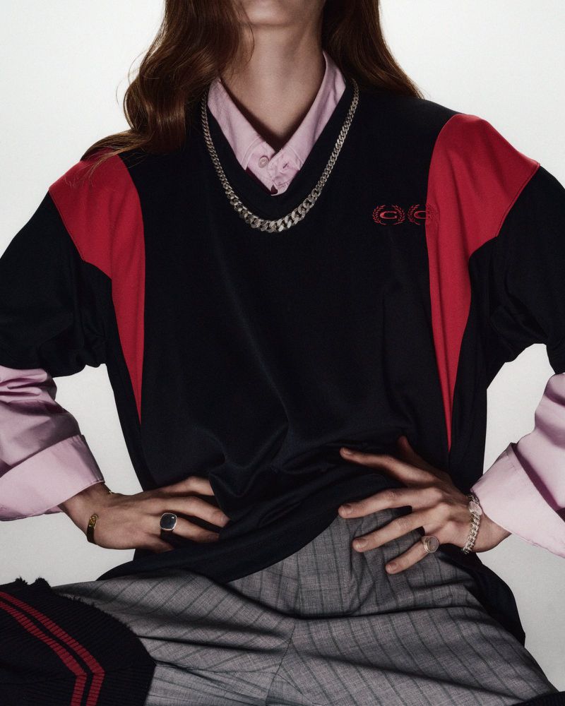Fashionable person in a black and red colorblock sweater with grey pinstripe pants and silver chain, posing indoors against a white background.
