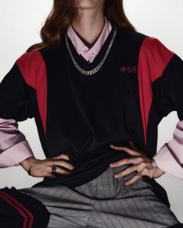 Model wearing a black and red v-neck jersey over a pink collared shirt with grey plaid trousers against a plain white background