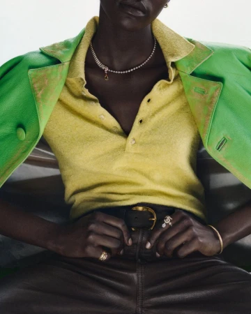 Close-up of a model wearing a green coat over a yellow polo shirt and dark leather trousers with a belt.
