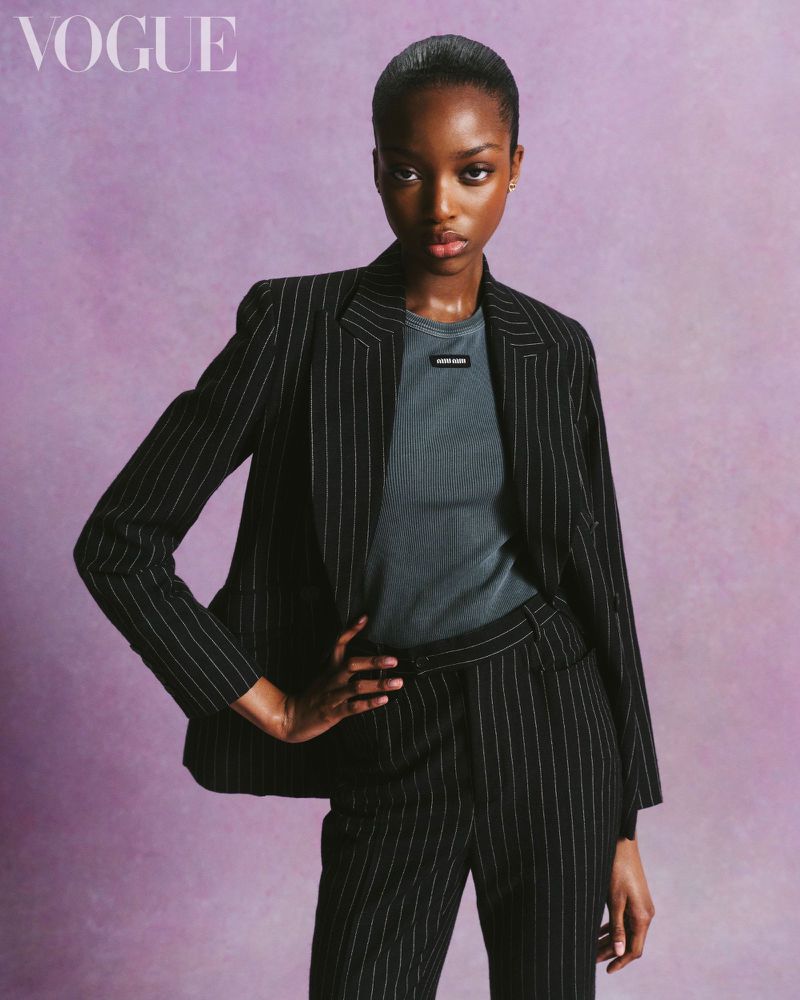 Elegant African American woman in a pinstripe suit poses against a muted purple backdrop for a fashion editorial, showcasing modern style.