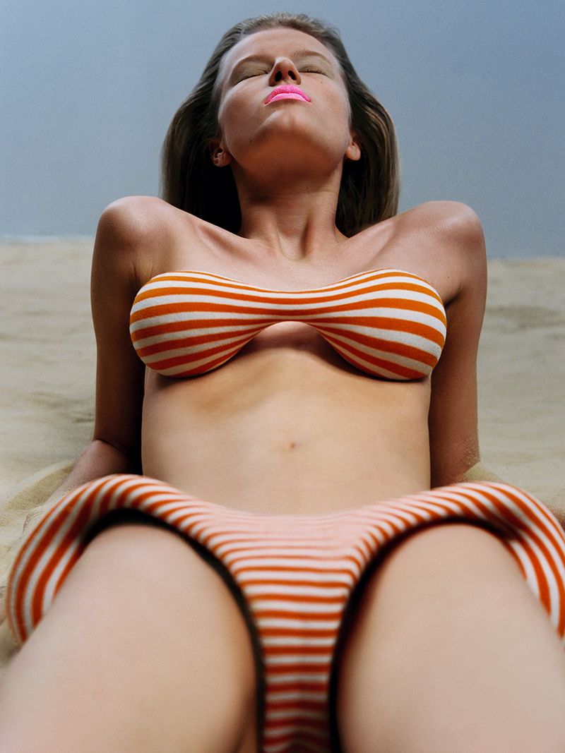 Woman with closed eyes lies on the sand in a striped orange and white swimsuit top and bottom enjoying the sunshine and warm weather.