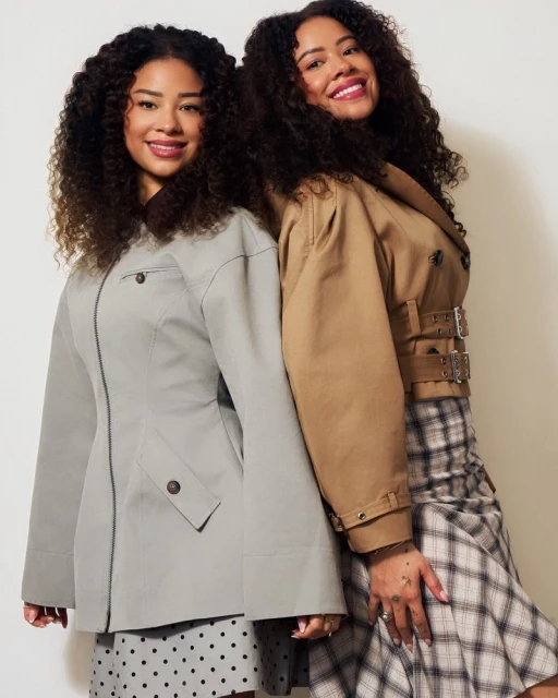 Two adult females with curly hair pose back to back indoors. One wears a beige trench coat with a plaid skirt, the other wears a grey coat.