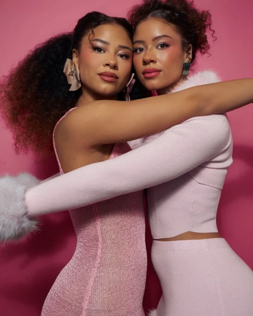 Two women with curly hair pose closely in pink outfits in front of a pink background in a studio setting with diffused lighting.