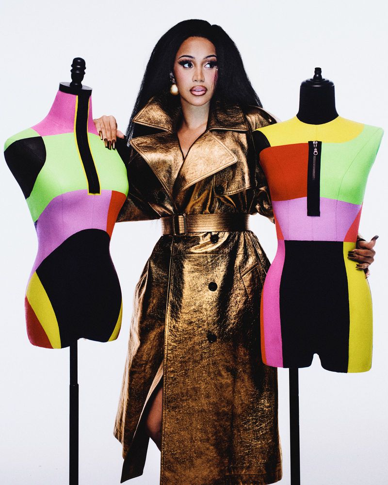 Portrait of adult female celebrity wearing metallic trench coat with black hair posing with colourful mannequins in a white studio setting.
