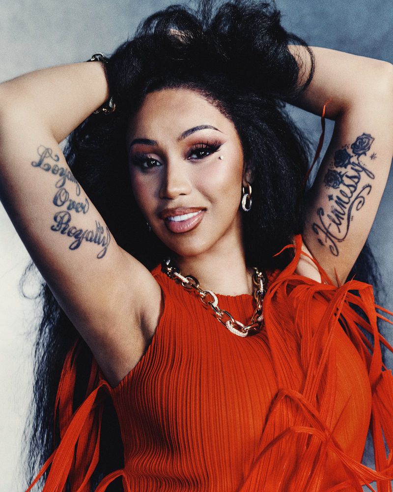 Close-up portrait of a smiling adult woman with dark hair, wearing a red top and gold chain, showcasing arm tattoos in a studio setting.