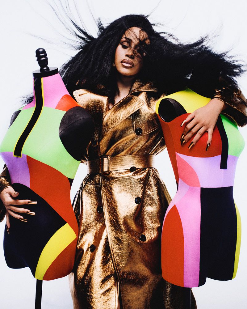 A woman in a metallic trench coat poses with colorful mannequins against a white backdrop in a fashion forward studio shot.