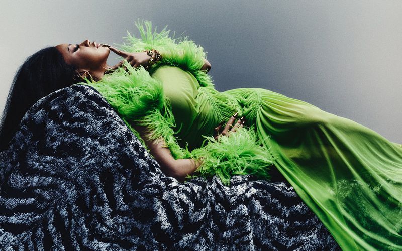 A woman with dark hair wearing a green dress with feather trim and gold bracelets, is lying on a patterned cushion, and gazing upward.