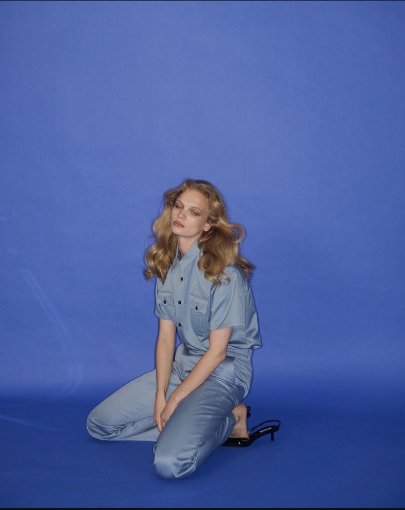 Woman in blue short-sleeve button-down shirt and pants kneeling on blue backdrop in studio. Black heeled sandals visible.