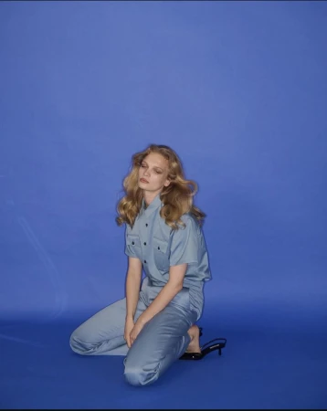 Woman in blue short-sleeve button-down shirt and pants kneeling on blue backdrop in studio. Black heeled sandals visible.