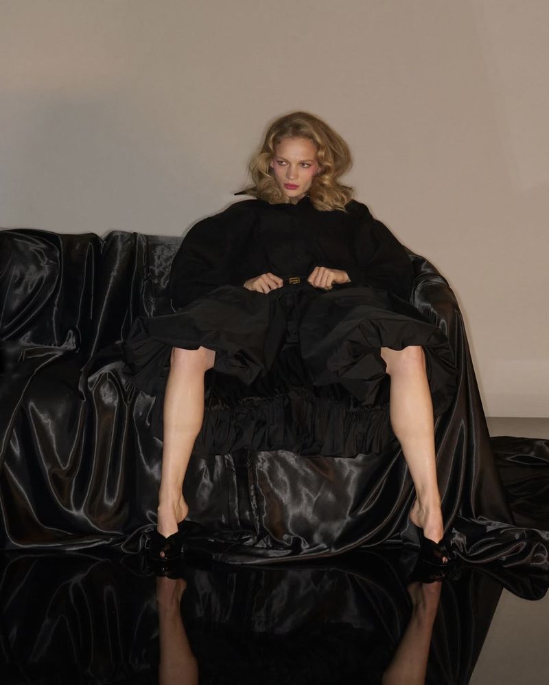 A woman with blonde wavy hair sits on a draped satin couch wearing a black dress and heels, posed in a photo studio with neutral background.