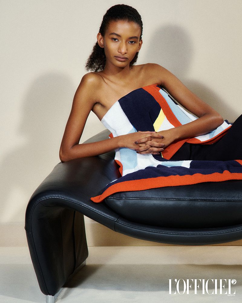 A young, dark-skinned woman with natural hair poses on a black chaise lounge, wearing a colorful strapless top against a neutral background.