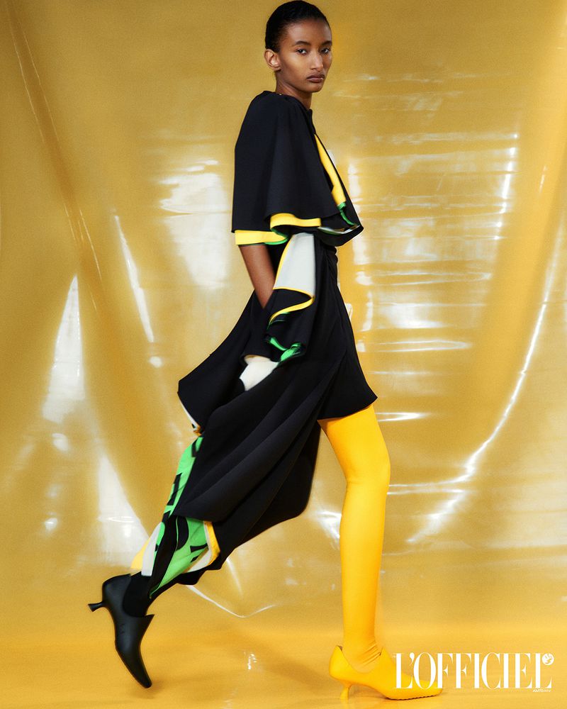 Fashion shot of a young Black woman in a black dress with a cape and yellow tights and shoes, posing against a yellow backdrop.