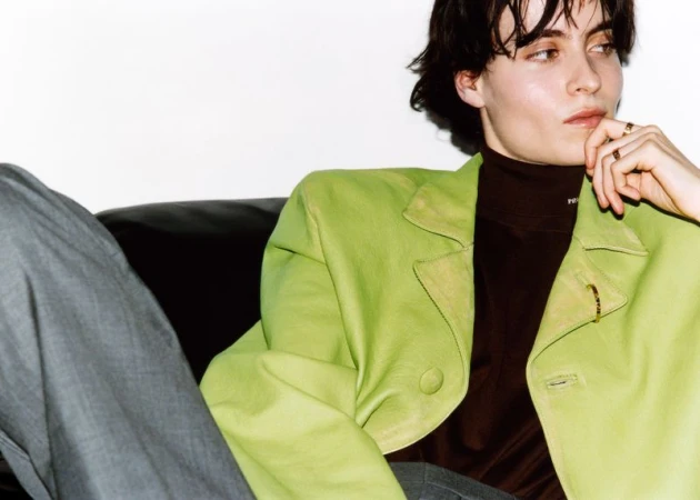 Portrait of a person with short dark hair wearing a lime-green blazer and brown turtleneck sitting on a black leather couch.