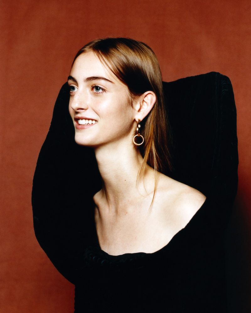 Smiling woman with light brown hair and blue eyes wearing gold hoop earrings against a dark brown background in a studio setting.