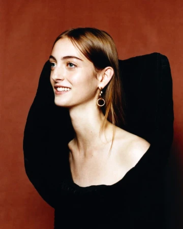 Smiling woman with light brown hair and blue eyes wearing gold hoop earrings against a dark brown background in a studio setting.