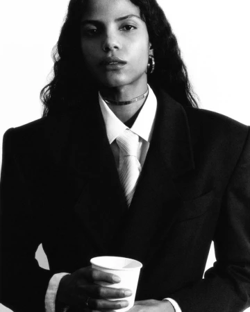 Black and white portrait of a person in a dark suit holding a paper cup against a stark white background, classic editorial photography.